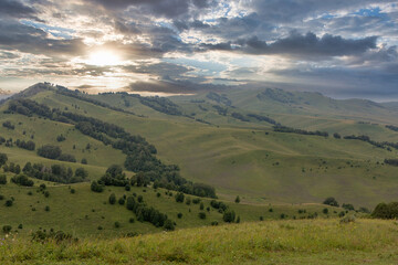 Obraz premium Wide panoramic view of Chyortov palets mountain with rolling green hills, meadow and dramatic cloudy sky at sunset in Altai Republic, Russia, tranquil wilderness scenery