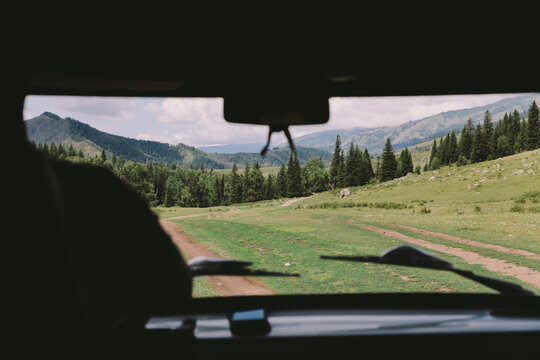 View from inside a vehicle of a winding dirt road across green meadows and pine forest in Ak-Kem Valley, Altai Republic, Russia, under a cloudy summer sky