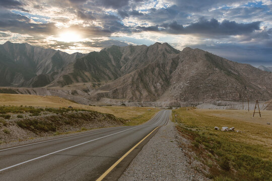 Empty asphalt road of the Chuysky Trakt stretching through rugged Altai Mountains at sunset, dramatic clouds and wide valley landscape, Altai Republic, Russia