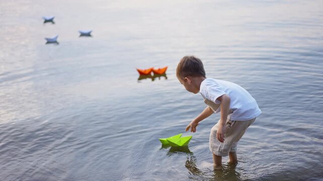 Elevated view of boy launching green paper boat in lake. Child bends over shallow water with colorful origami fleet floating nearby.