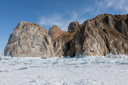 Frozen Lake Baikal coastline at Cape Three Brothers with rugged granite cliffs, blue sky, and cracked drift ice on the shore in Irkutsk Oblast, Russia