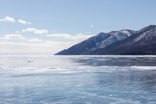 Wide winter landscape of frozen Lake Baikal with smooth blue ice, snow-covered mountains and clear sky at Peschanaya Bay, Irkutsk Oblast, Russia