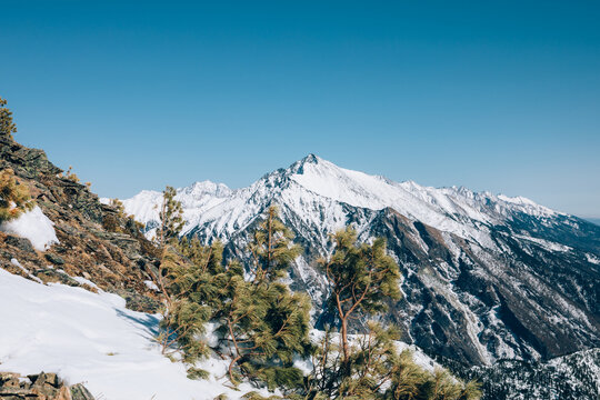 Wide winter landscape view of Peak Galina near Arshan, Republic of Buryatia, Russia, with snow-covered mountain ridges, rocky slopes, pine trees and clear blue sky
