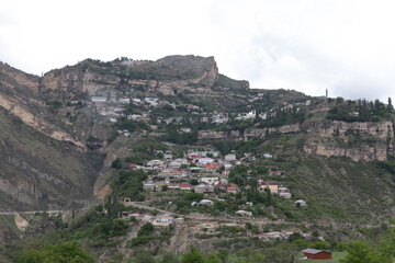 Obraz premium Wide panoramic view of Gunib aul built on steep rocky slopes and terraces, with houses and greenery under cloudy sky, Dagestan, Russia, Caucasus Mountains
