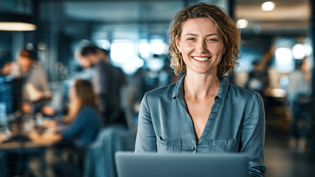 Smiling businesswoman working with a laptop in a busy modern office, confident professional surrounded by an active workplace environment with colleagues collaborating in the background, 