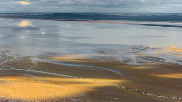 Aerial view of vast salt flats and a reflective lake under a cloudy sky with golden light hitting the shoreline in Aktau, Mangystau Region, Kazakhstan.