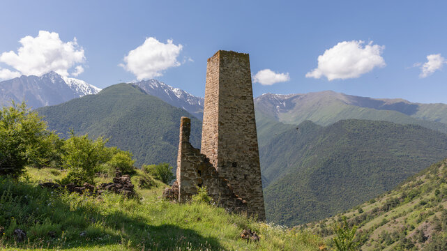 Scenic view of the medieval Erzi tower complex ruins on a green mountain ridge with high peaks and clouds, Dzheyrakhsky District, Republic of Ingushetia, Russia