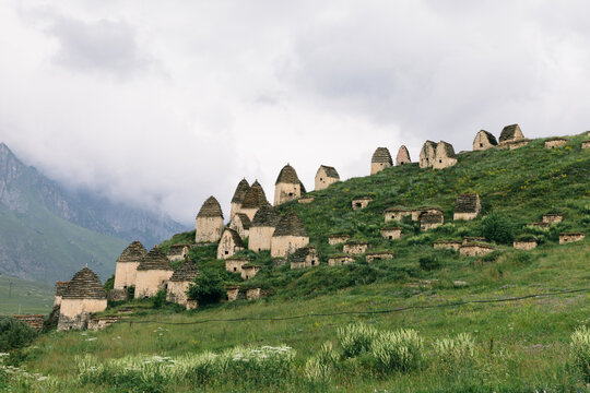 Wide view of the Dargavs City of the Dead necropolis with traditional stone crypts on a green mountain slope in North Ossetia&ndash;Alania, Russia under cloudy sky