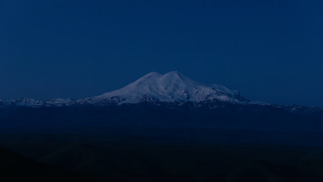 Distant view of snowcapped Mount Elbrus at blue hour, seen from Bermamyt Plateau in Karachay-Cherkessia, Russia, with dark ridgelines and clear sky
