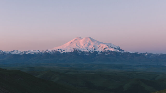 Panoramic sunrise view of snowcapped Mount Elbrus from Bermamyt Plateau, Karachay-Cherkess Republic, Russia, with soft pink sky and layered mountain ridges