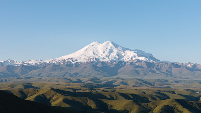 Scenic view of snowcapped Mount Elbrus from Bermamyt Plateau, Karachay-Cherkess Republic, Russia, with rolling green hills and clear blue sky in daylight
