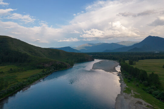 Aerial landscape of a winding river through forest and meadow with distant mountain ridges under dramatic clouds near Zamulta, Altai Republic, Russia at dusk