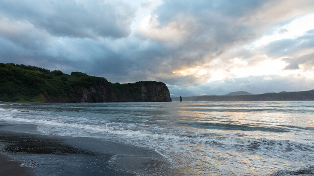Dramatic cloudy sky over a dark volcanic sand beach with gentle waves and rugged sea cliffs, photographed on the Kamchatka Peninsula, Russia, at dusk