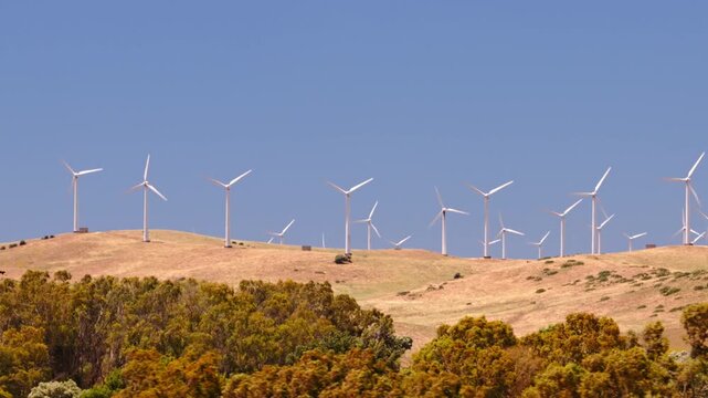 Cinematic drone footage of a large wind farm with turbines generating clean renewable electricity across a rural landscape. Sustainable energy infrastructure supporting decarbonization and climate goa