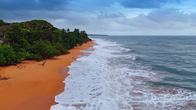 Drone view of a wild red sand beach with jungle, Bocas del Toro, Panama. Diagonal coastline of ochre sand, white surf and dense green canopy under dramatic grey clouds.