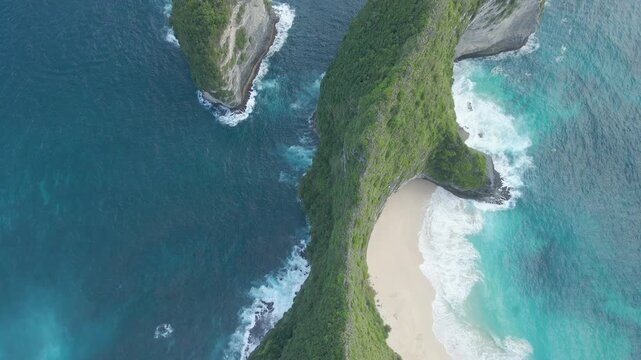 Cinematic aerial shot of the iconic Kelingking Beach and TRex shaped cliff in Nusa Penida, Bali. High angle view of turquoise ocean waves crashing against white sand and limestone rocks.