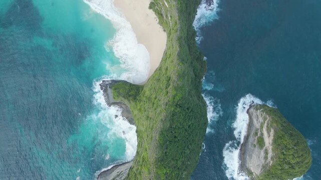 Cinematic aerial topdown perspective of the iconic Kelingking Beach cliff in Nusa Penida, Bali. Features turquoise waves crashing against white sand and lush green limestone formations.