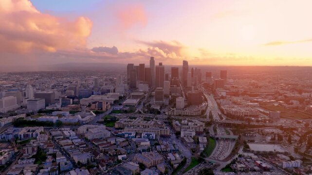 Aerial view of downtown Los Angeles shows traffic congestion during rush hour. Skyscrapers outline the skyline as sunset lights up the city, highlighting the urban infrastructure and palm trees.