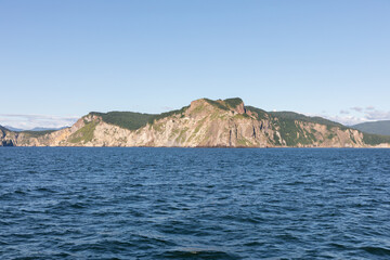 Naklejka premium Wide seascape with rocky coastal cliffs and green headland under clear blue sky, viewed from the water in Kamchatka Krai, Russia, on the Pacific Ocean