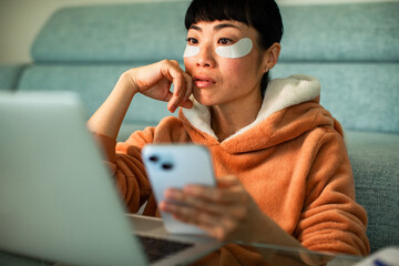 Woman with under-eye patches using laptop and phone at home © Geber86
