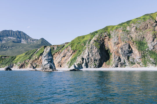 Scenic seascape with rugged volcanic cliffs, green slopes, and calm blue water under clear sky on the Kamchatka Peninsula, Kamchatka Krai, Russia