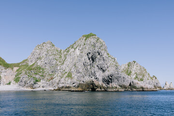 Naklejka premium Scenic view of rugged coastal cliffs with green vegetation rising above calm blue sea under a clear sky on the Kamchatka Peninsula, Kamchatka Krai, Russia