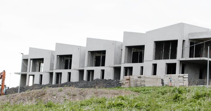 A row of modern, unfinished concrete houses on a construction site with mounds of dirt and weeds in the foreground under an overcast sky.