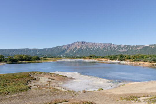 Wide view of a calm lake inside Uzon Volcano caldera with mineral shores, tundra vegetation and mountain ridge under clear sky, Kamchatka Krai, Russia