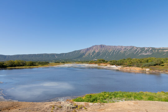 Wide scenic view of Uzon Volcano caldera with calm lake, geothermal shoreline and green tundra under clear blue sky, Kamchatka Krai, Russia