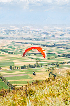 Vertical shot of a tandem paraglider with a bright red canopy flying over agricultural fields in Karakol, Kyrgyzstan.