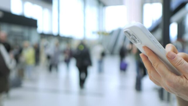 Hand holds smartphone in busy airport terminal. Blurred travelers walk with luggage behind. Bright daylight shines through large windows. Focus remains on phone and hand only