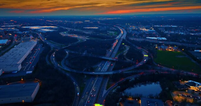 Aerial view of a road interchange next to a pond, United States. High viewpoint showing still water reflecting the sky, complex looping ramps, moving vehicles, and a colorful evening horizon.