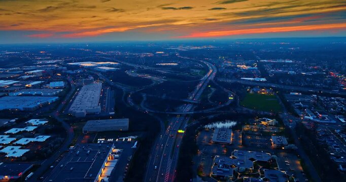 Panoramic view of a highway junction near warehouses, United States. High drone angle capturing bright orange twilight sky, vast industrial complexes, and intricate networks of interlocking ramps.