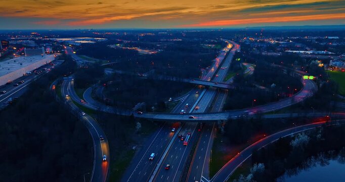 Sunset sky over a curved highway cloverleaf ramp, United States. Elevated drone shot showing a circular asphalt turnoff over main lanes, moving cars, dense dark trees, and vibrant evening light.