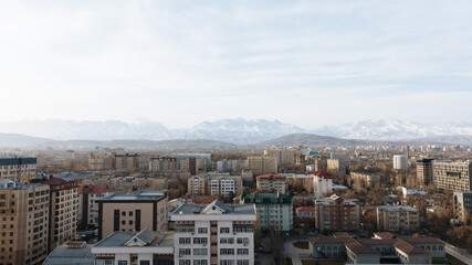 Fototapeta premium Wide urban skyline view of Bishkek, Chuy Region, Kyrgyzstan, with residential buildings and distant Tien Shan mountain range under soft daylight and clouds