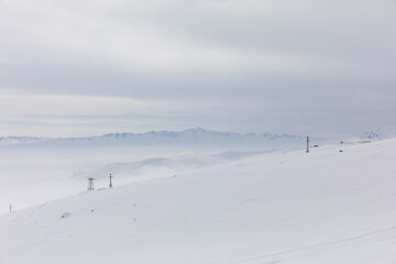 Fototapeta premium Wide winter view of Ala-Bel Pass with fresh snowdrifts, low fog and distant mountain ridge under an overcast sky, Kyrgyzstan, Central Asia travel scenery