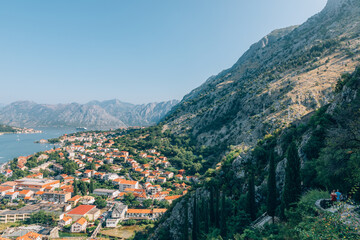 Obraz premium Wide panoramic landscape of Kotor and the Bay of Kotor with red tiled rooftops, Adriatic coastline and rugged mountains under clear blue sky, Montenegro