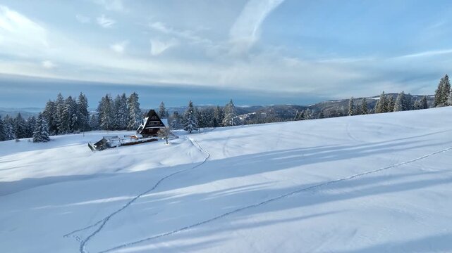 Aerial forward flight over snow covered mountain meadow towards wooden chalet along footprint path on sunny winter day