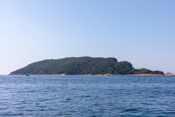 Fototapeta premium Wide view of Sveti Nikola Island with green hills and rocky shore across calm Adriatic Sea water near Budva, Montenegro, under clear blue sky on a sunny day