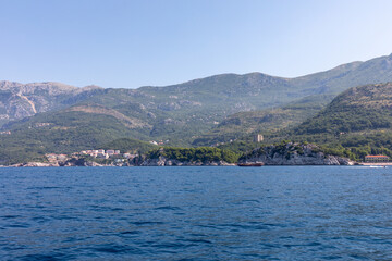 Naklejka premium Wide seascape with calm Adriatic waters, rocky coastline and green mountains near Budva, Coastal Region, Montenegro, with a small boat offshore under blue sky
