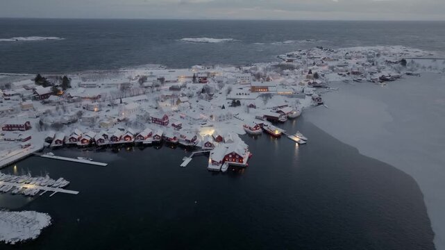 Aerial dusk view of a snow-covered coastal fishing village with red boathouses and frozen harbor