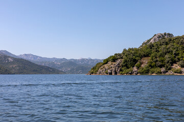 Naklejka premium Wide landscape view of Lake Skadar with calm blue water, rocky green hills and distant mountains under clear sky, Skadar Lake National Park, Montenegro