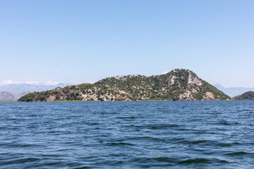 Naklejka premium Wide panoramic view of a rocky green island and rippling water on Lake Skadar, Montenegro, with distant mountains under a clear blue sky on a sunny day