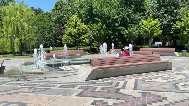 Park Fountain Oasis: A refreshing fountain sprays water in a vibrant urban park, offering a cool respite on a sunny day. People relax on benches, enjoying the tranquil atmosphere and lush greenery.