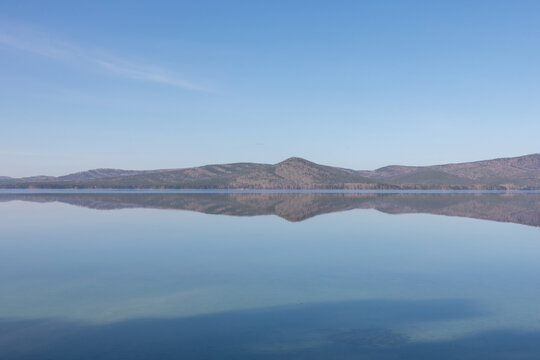 Serene landscape of Lake Turgoyak with still water creating a perfect mirror reflection of distant mountains under a clear blue sky, peaceful nature background