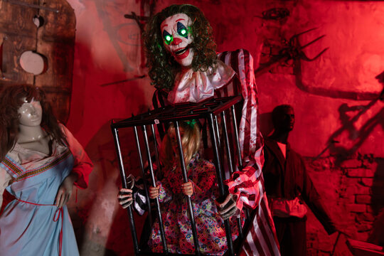 Satka, Russia - May 9, 2025: Scary Halloween attraction scene at Sonkina Lagoon, Satka, Russia with creepy clown performer and child behind metal bars, red lighting and haunted house decor