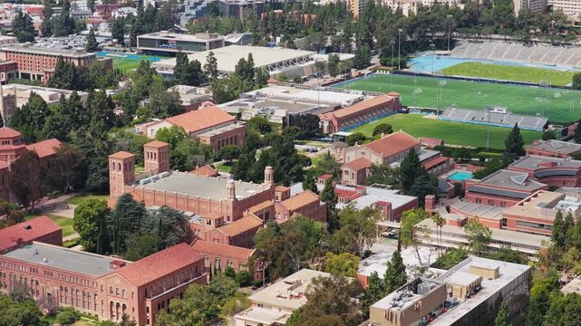 Aerial shot flying over University of California Los Angeles campus with red brick buildings and sports fields on clear summer day in suburban neighborhood