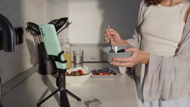 Young adult Caucasian female influencer recording healthy breakfast preparation on smartphone mounted on tripod. Video is suitable for social media marketing and healthy lifestyle campaigns