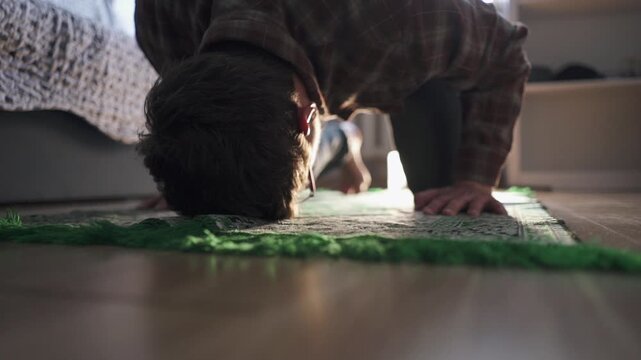 Muslim man performing prostration Sujood on a green prayer rug at home, bowing down in prayer.