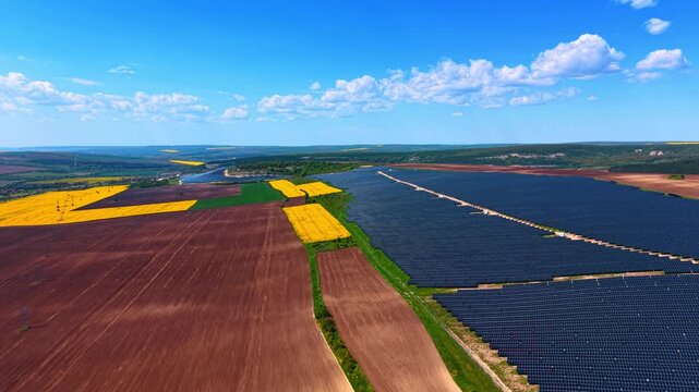 Colorful farmland mosaic with solar panels and rapeseed patches. Elevated drone shot showing dark blue arrays, yellow canola, red earth, and green fields under scattered clouds.